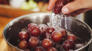 A close-up of a hand rinsing Muscadine grapes in a metal bowl, with water droplets splashing around