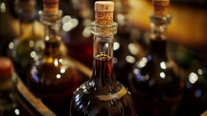 A close-up of a glass bottle filled with dark muscadine wine, sealed with a cork, surrounded by other wine bottles in soft lighting