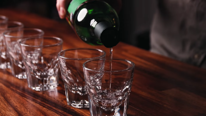 A Person Pouring Mezcal from A Bottle Into Shot Glasses on A Wooden Surface