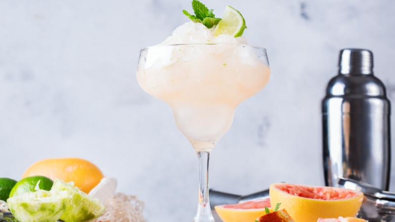 A mezcal cocktail served over crushed ice in a margarita glass, garnished with lime and mint, surrounded by fresh citrus and a shaker