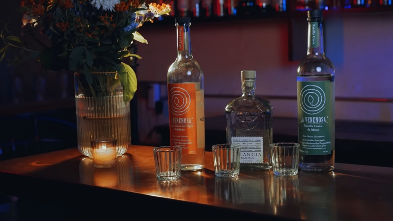 Bottles of mezcal and raicilla with shot glasses on a bar beside a vase of flowers and a lit candle