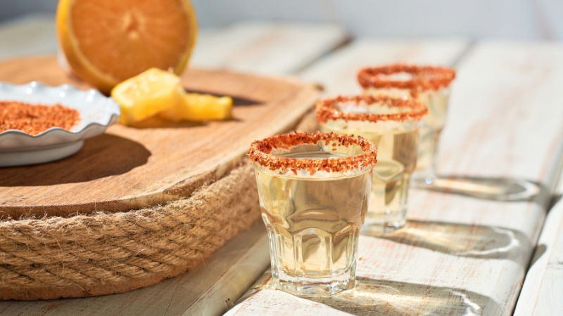 Three shot glasses with chili-rimmed edges filled with agave spirit, served alongside orange and pineapple slices