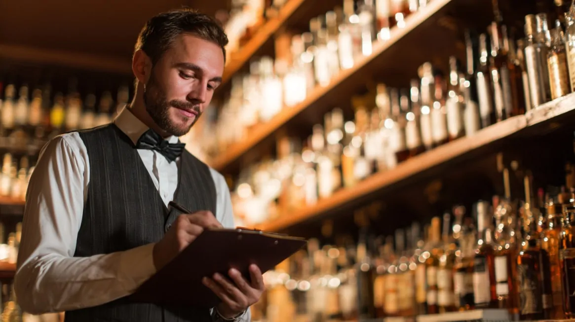 A bartender in formal attire writes on a clipboard while standing in front of shelves filled with liquor bottles